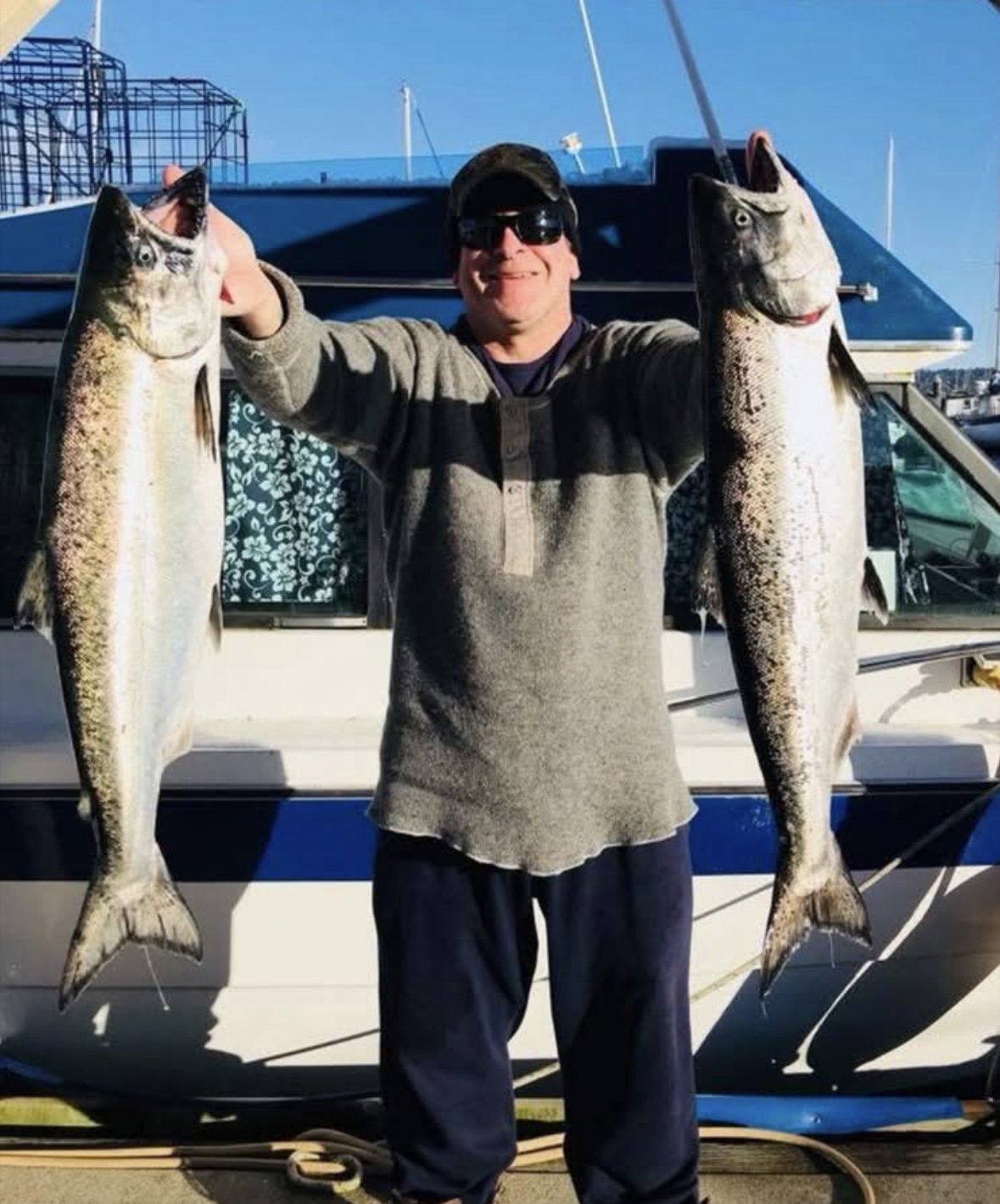 Douglas smiling and presenting two fish he caught to the camera on a dock. Behind him is a boat and sunny skies.